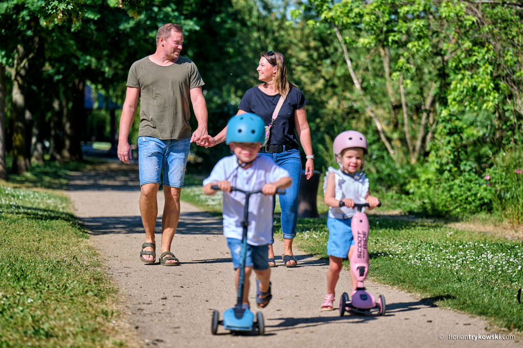 Eine Familie geht Spazieren im Grünen. Im Vordergrund zwei Kinder mit Tretrollern, dahinter die Eltern Hand in Hand.